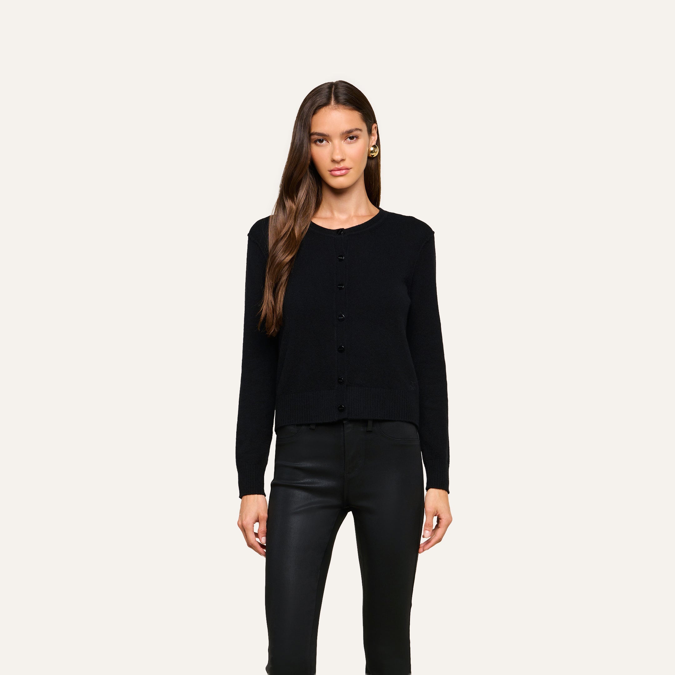 A woman with long brown hair stands against a plain off-white background, facing forward with a neutral expression, wearing the black Malone Cashmere Cardigan and black leather pants.
