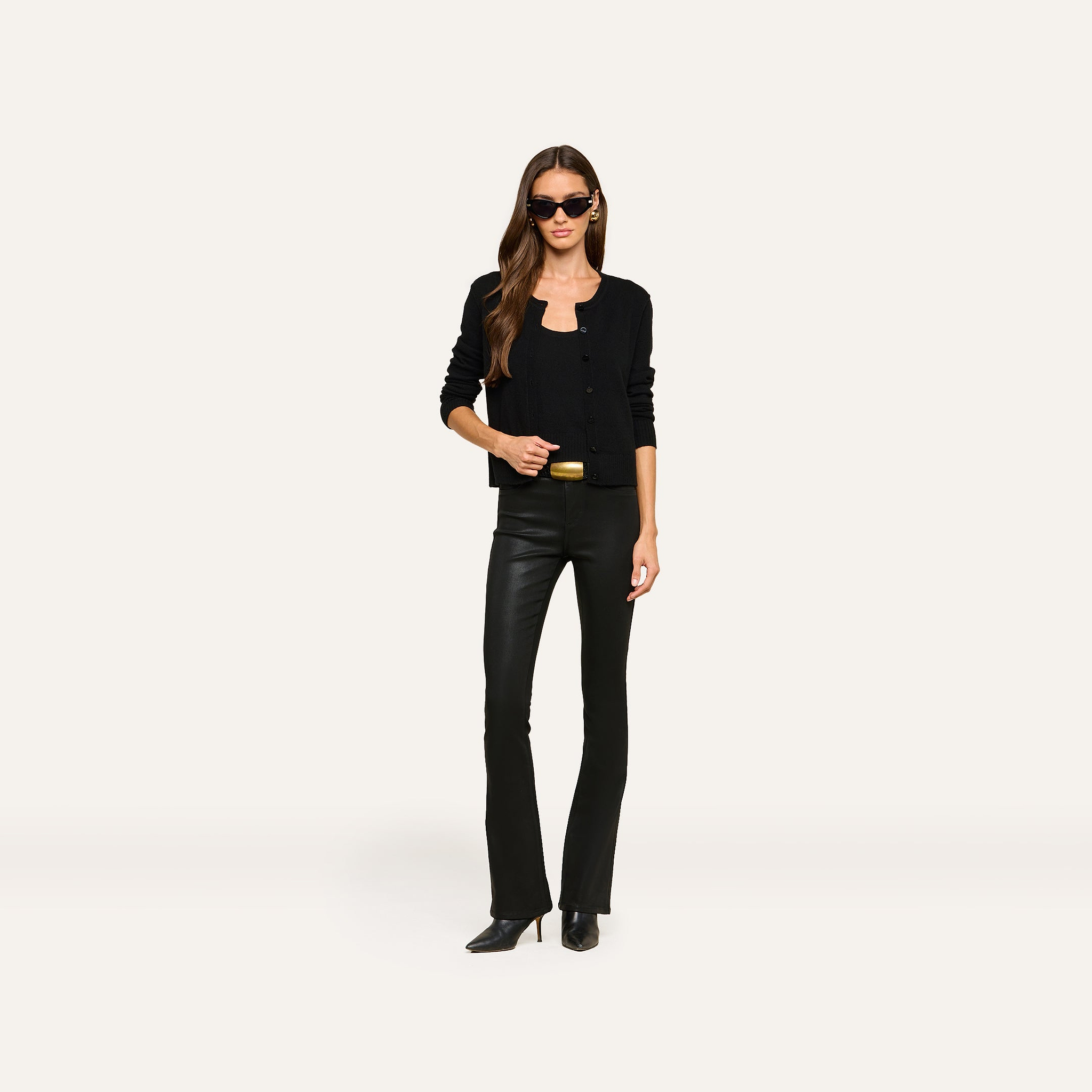 A woman wearing sunglasses, the Malone Cashmere Cardigan in black, black flared leather pants, and black shoes stands against a plain white background.