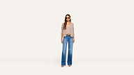 A woman wearing sunglasses, a beige cardigan, stylish blue wide-leg jeans, and brown shoes stands against a plain white background.