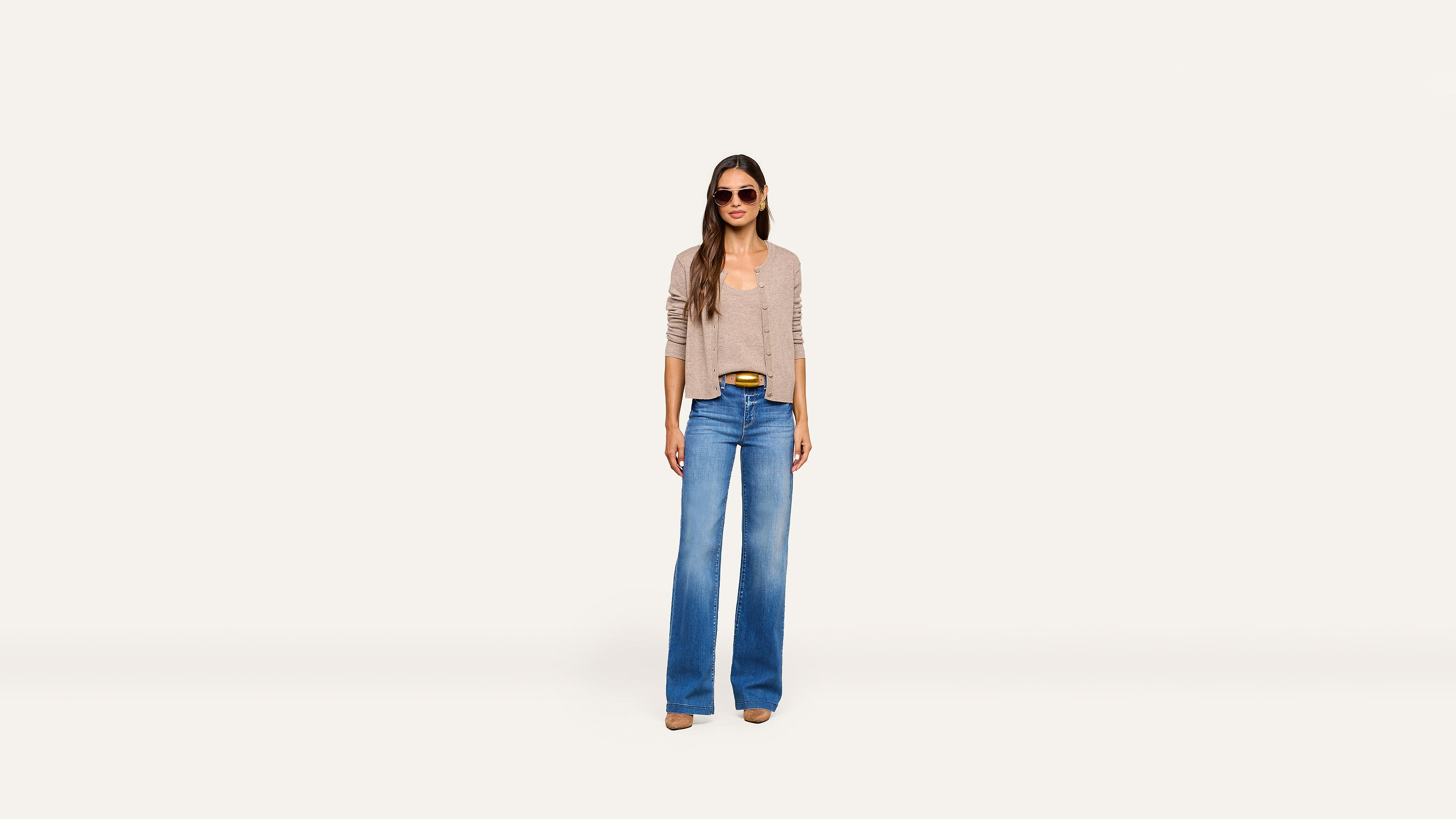 A woman wearing sunglasses, a beige cardigan, stylish blue wide-leg jeans, and brown shoes stands against a plain white background.