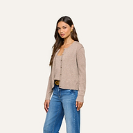 A woman with long brown hair and gold hoop earrings stands against a plain light background, wearing the Malone Cashmere Cardigan in beige over a matching top and blue jeans. She has a neutral expression.