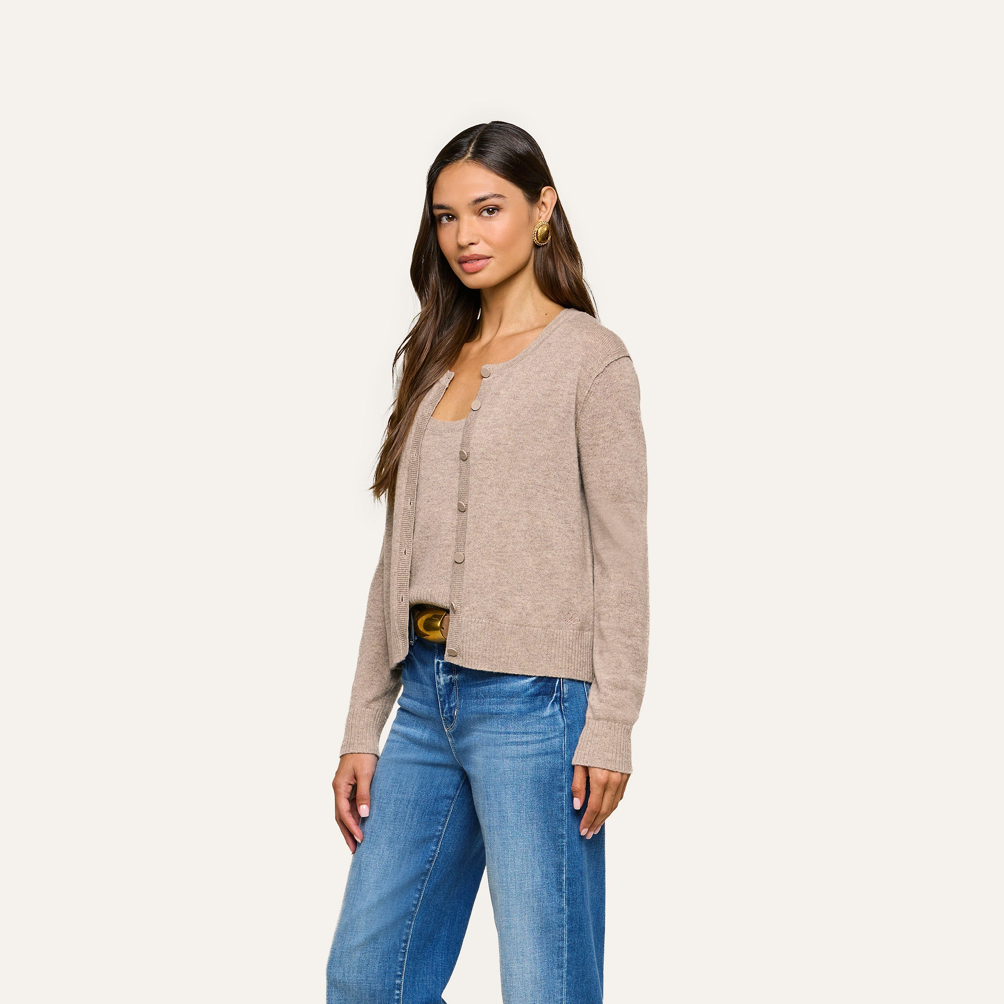 A woman with long brown hair and gold hoop earrings stands against a plain light background, wearing the Malone Cashmere Cardigan in beige over a matching top and blue jeans. She has a neutral expression.