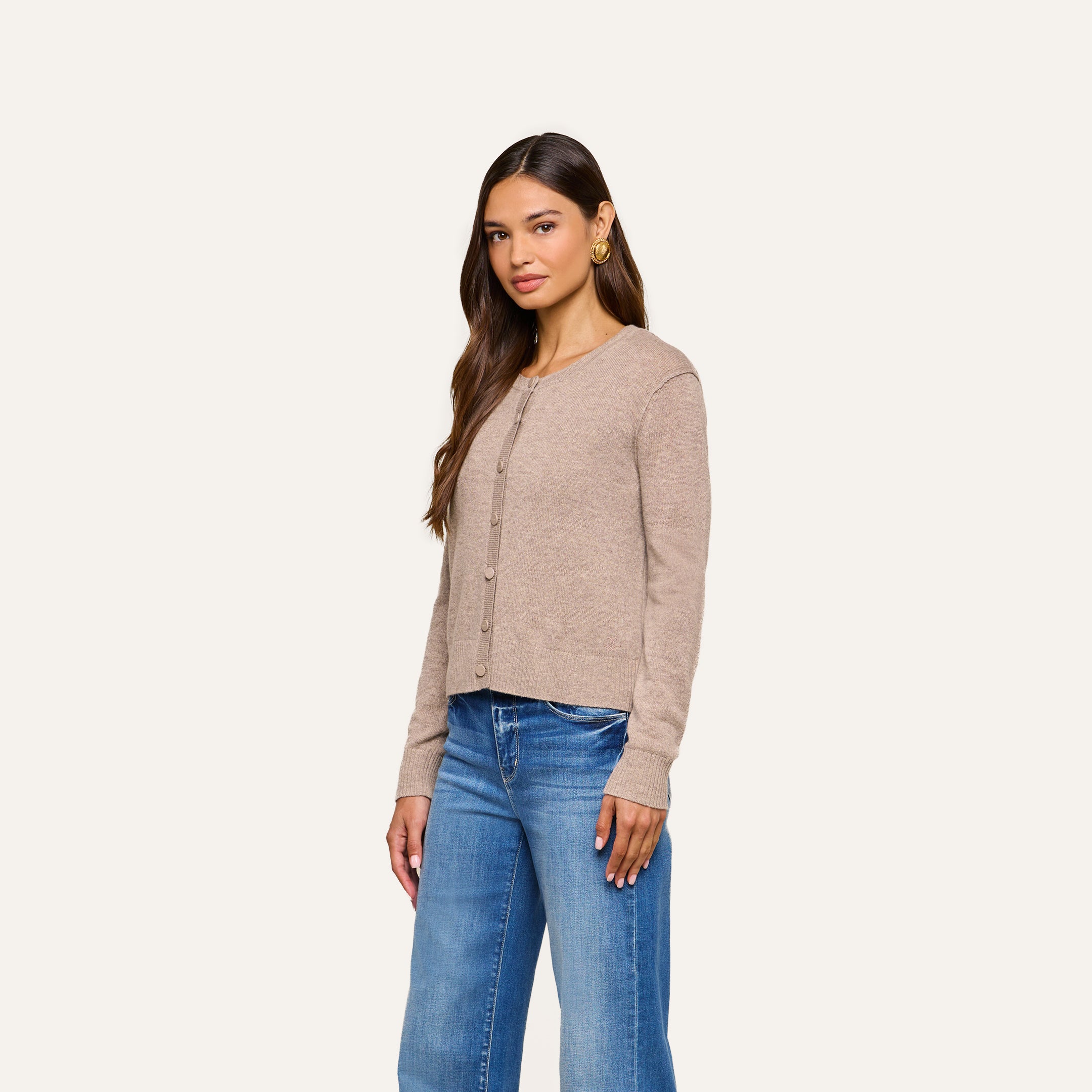 A woman wearing the Malone Cashmere Cardigan and blue jeans stands against a plain off-white background, looking slightly to the side with a neutral expression.