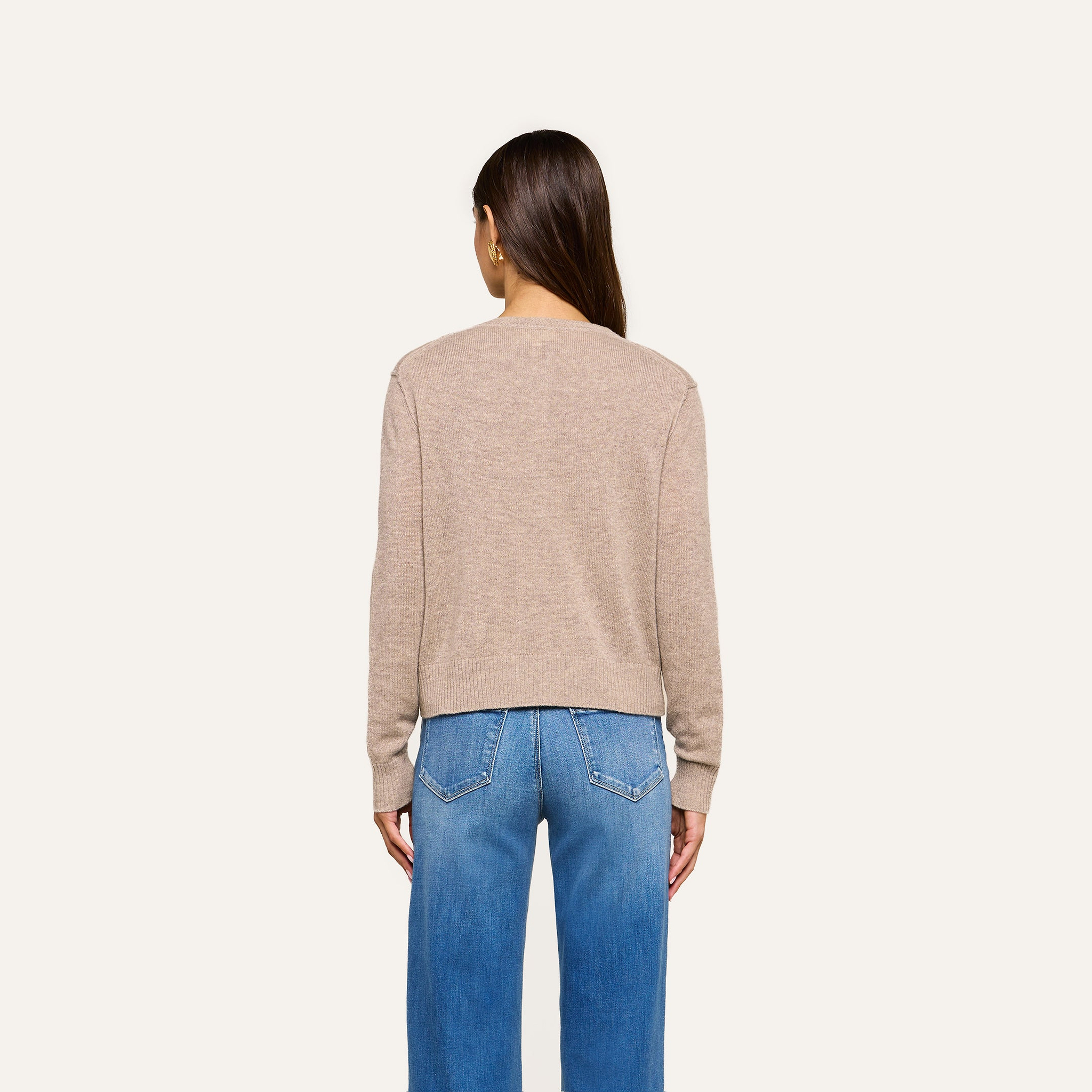 A person with long brown hair, wearing the Malone Cashmere Cardigan in light beige and blue jeans, stands facing away from the camera against a plain light background.