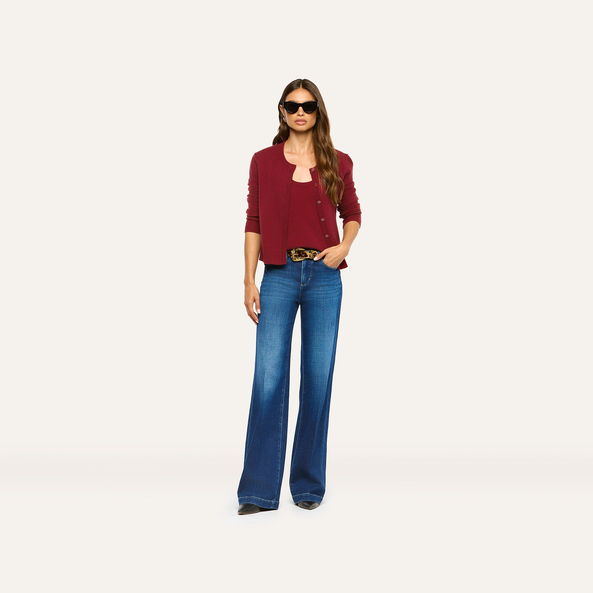 A person wearing a cherry red Malone Cashmere Cardigan over a matching tank top and flared blue jeans with a patterned belt.
