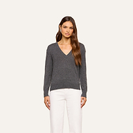 A woman with long brown hair is wearing the Spence Cashmere Pullover in dark gray along with white pants, standing against a plain light background. Her expression is neutral and her arms are relaxed at her sides.
