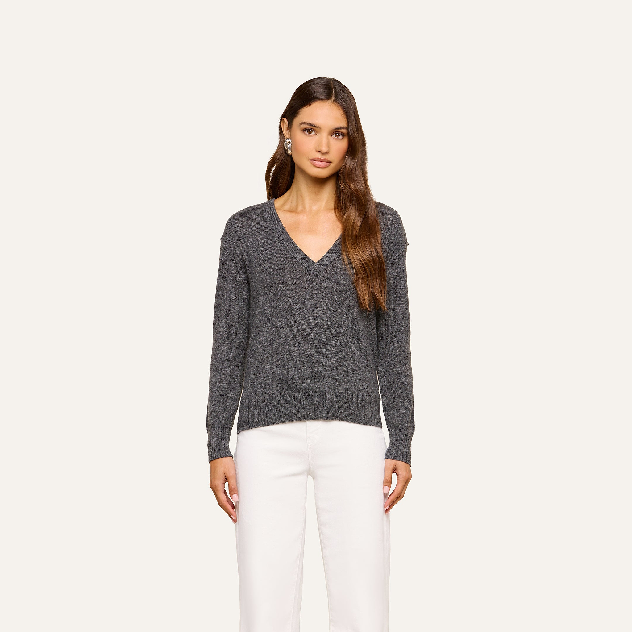 A woman with long brown hair is wearing the Spence Cashmere Pullover in dark gray along with white pants, standing against a plain light background. Her expression is neutral and her arms are relaxed at her sides.