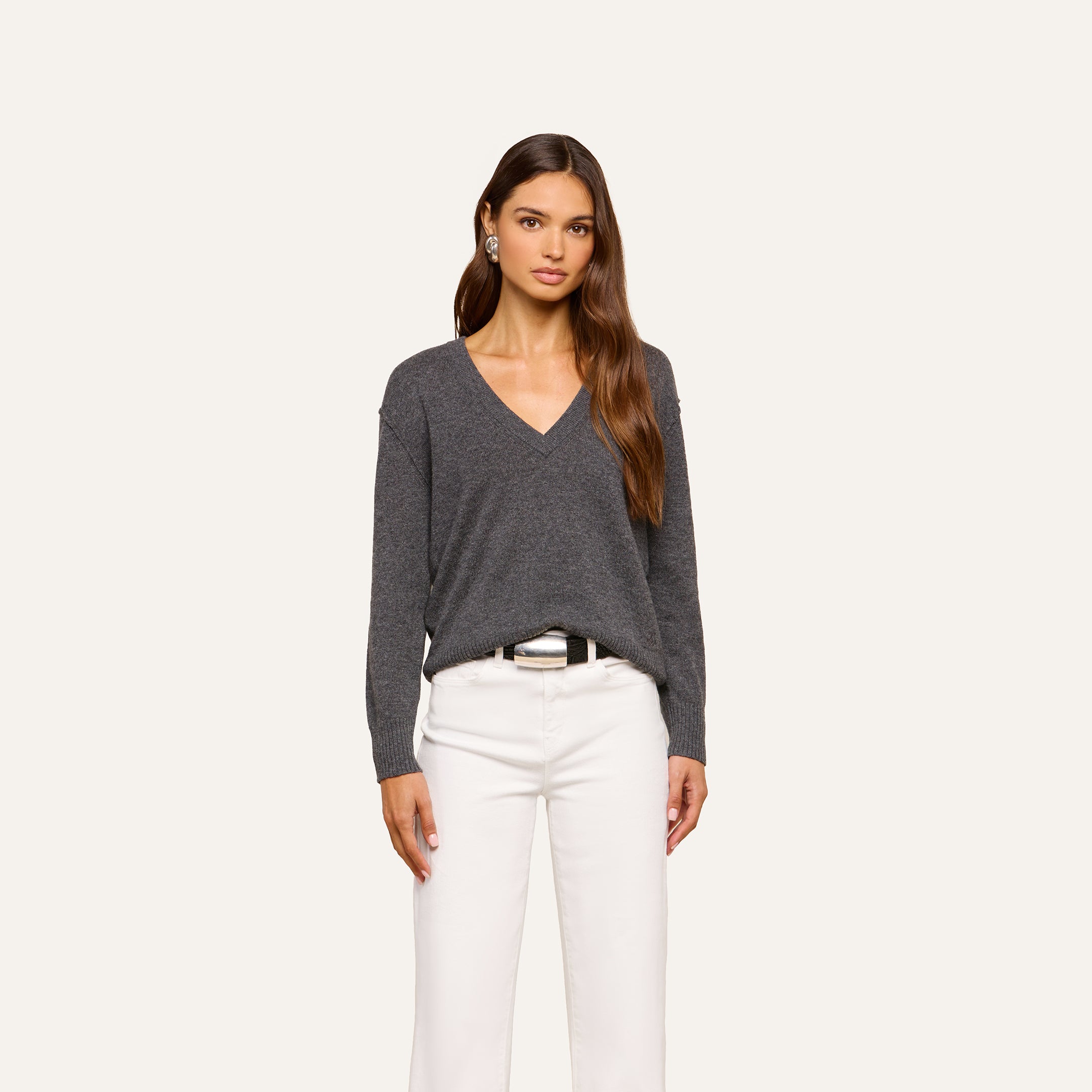 A woman with long brown hair wears the Spence Cashmere Pullover in dark gray and white pants, standing against a plain light background and looking at the camera with a neutral expression.
