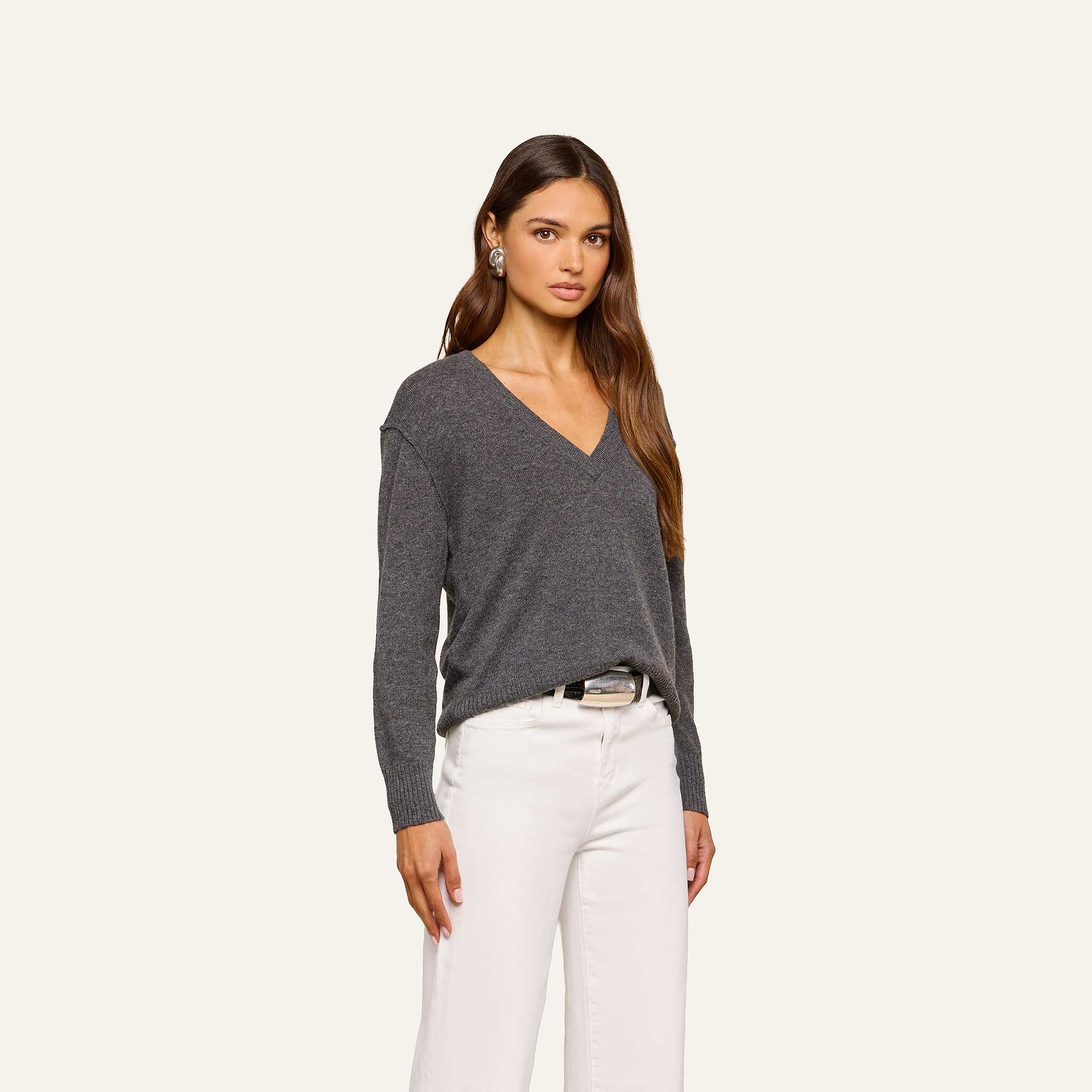 A woman with long brown hair wearing the Spence Cashmere Pullover—a women’s V-neck sweater in dark gray—paired with white pants, stands against a plain light background, looking at the camera with a neutral expression.