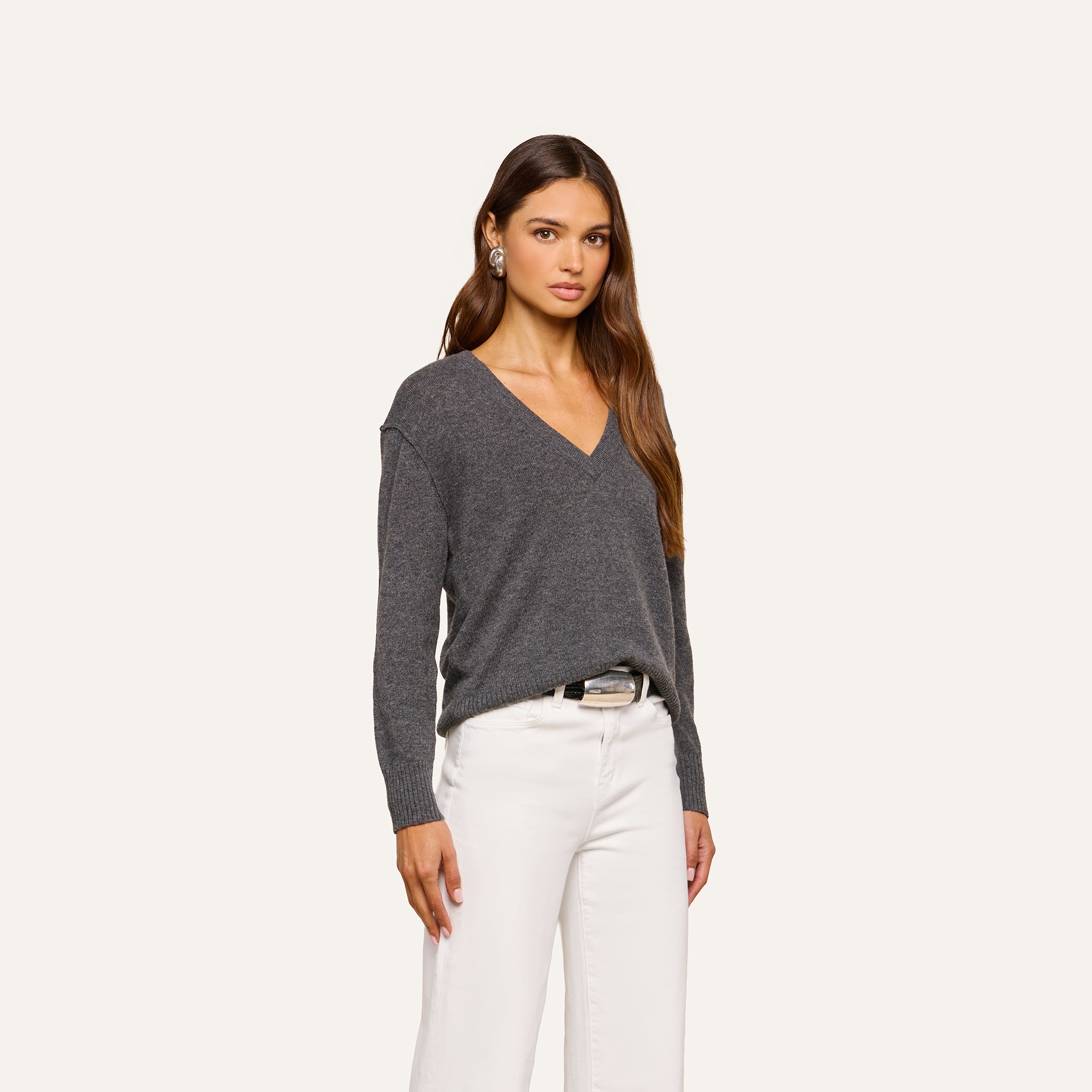 A woman with long brown hair wearing the Spence Cashmere Pullover—a women’s V-neck sweater in dark gray—paired with white pants, stands against a plain light background, looking at the camera with a neutral expression.