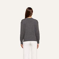 A person with long brown hair is shown from the back, wearing the Spence Cashmere Pullover—a lightweight women’s v-neck sweater in gray—paired with white pants against a plain, light background.