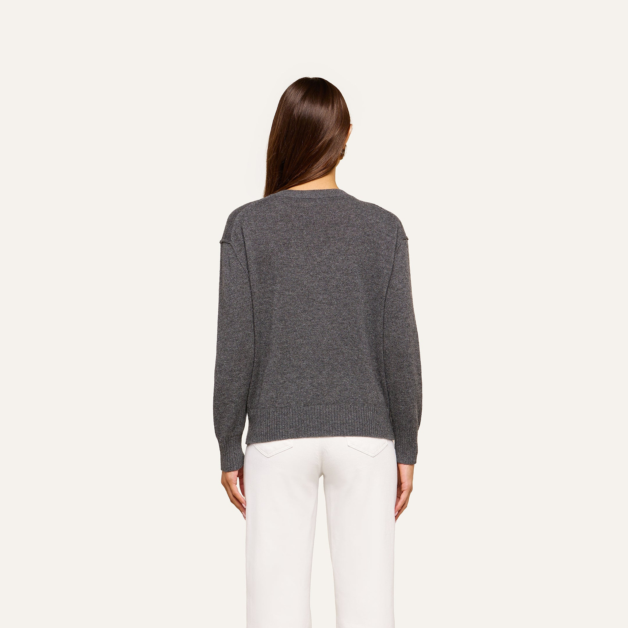 A person with long brown hair is shown from the back, wearing the Spence Cashmere Pullover—a lightweight women’s v-neck sweater in gray—paired with white pants against a plain, light background.