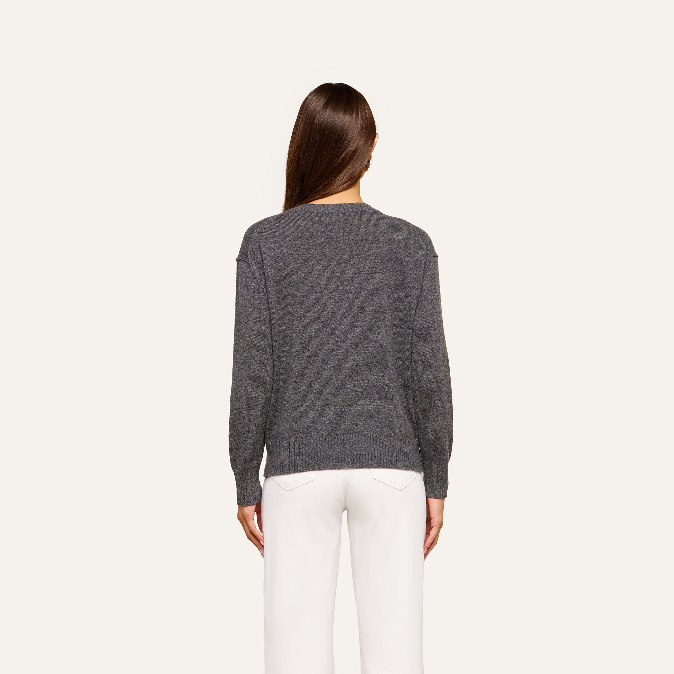 A person with long brown hair is shown from the back, wearing the Spence Cashmere Pullover—a lightweight women’s v-neck sweater in gray—paired with white pants against a plain, light background.