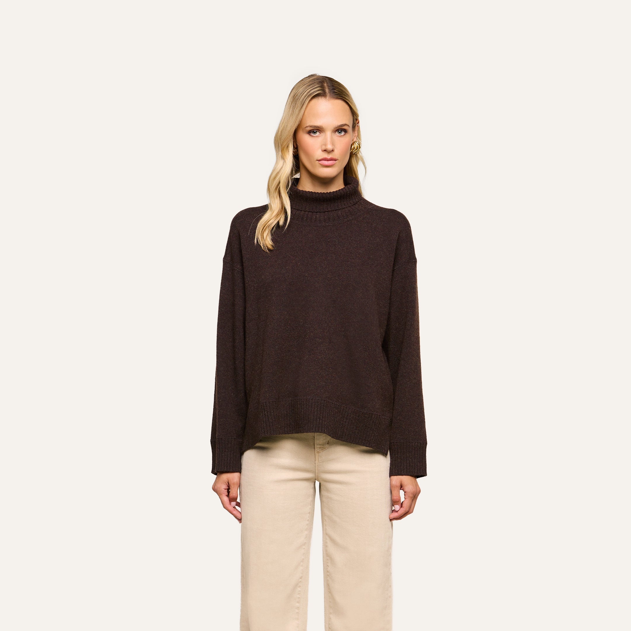 A woman with long blonde hair wears the Selwyn Oversized Turtleneck in dark brown, paired with light beige pants, standing against a plain light background.