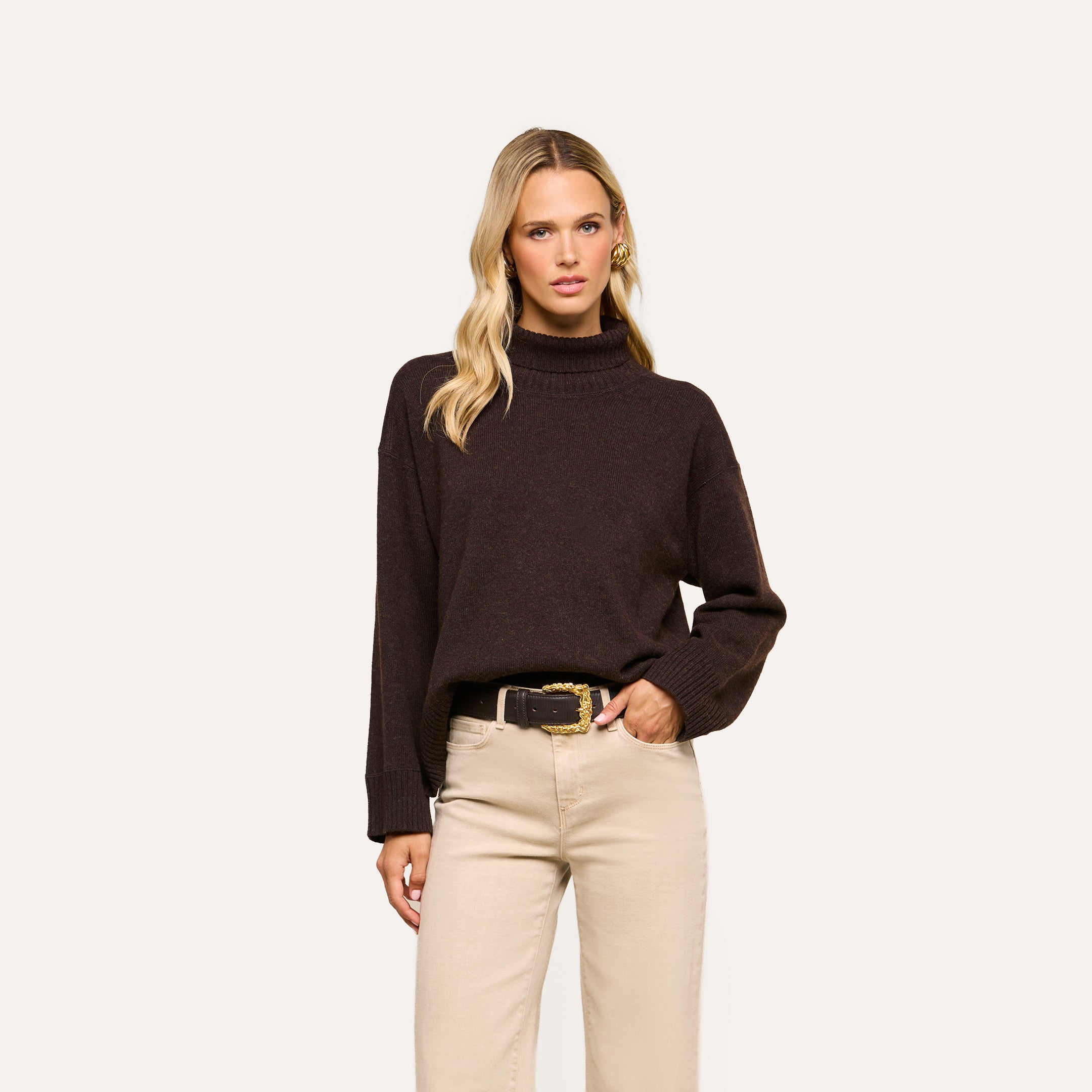 Wearing the Selwyn Oversized Turtleneck in dark brown, paired with beige pants and a black belt with a gold buckle, a woman stands against a plain light background and looks at the camera.