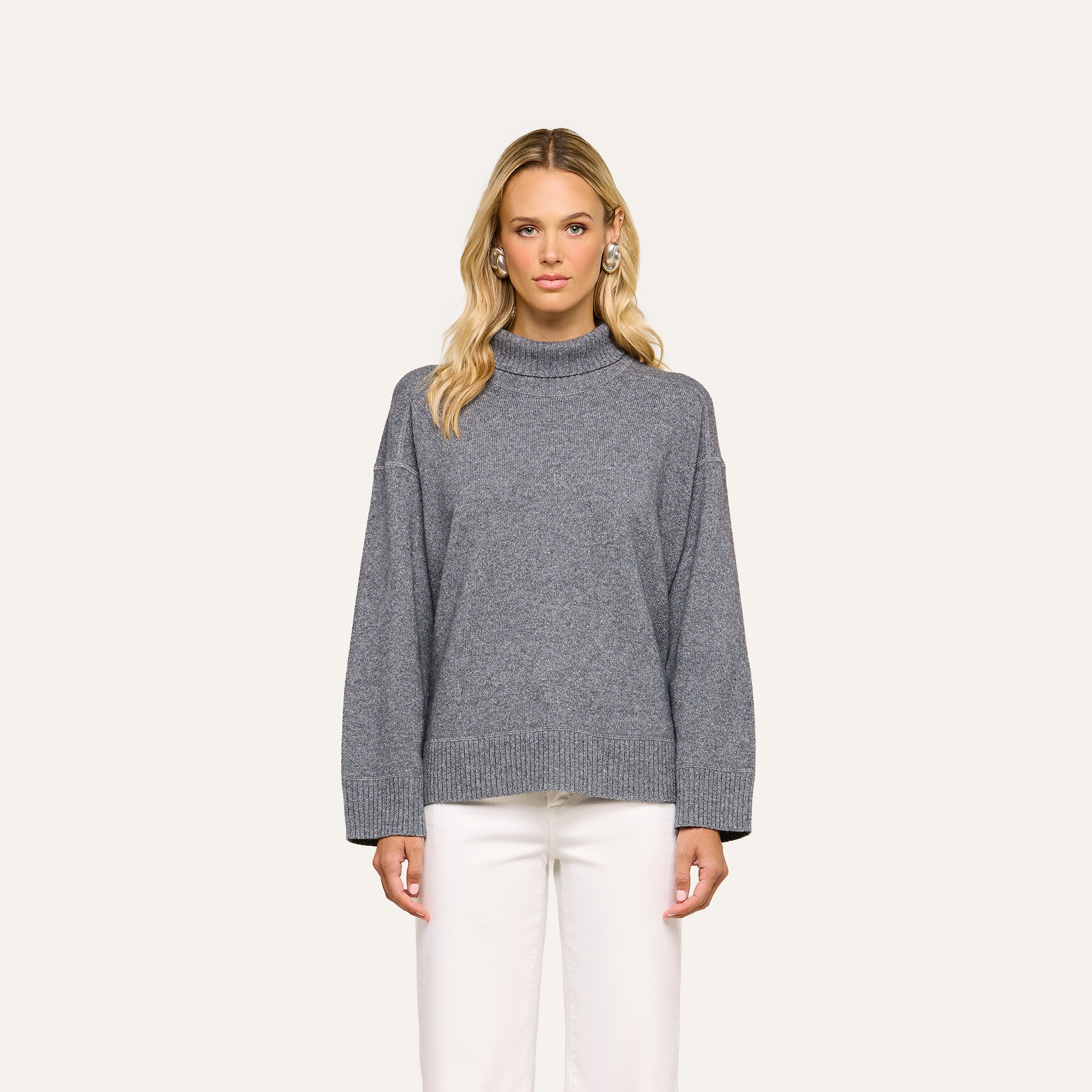 Woman with long blonde hair wearing the Selwyn Oversized Turtleneck in heather grey wool-cotton and white pants, standing against a plain light background, facing forward with a neutral expression.