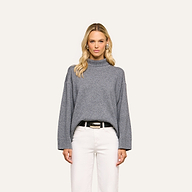 A woman with long blonde hair wears the Selwyn Oversized Turtleneck in heather grey paired with white pants, standing against a plain light background and looking straight at the camera with a neutral expression.