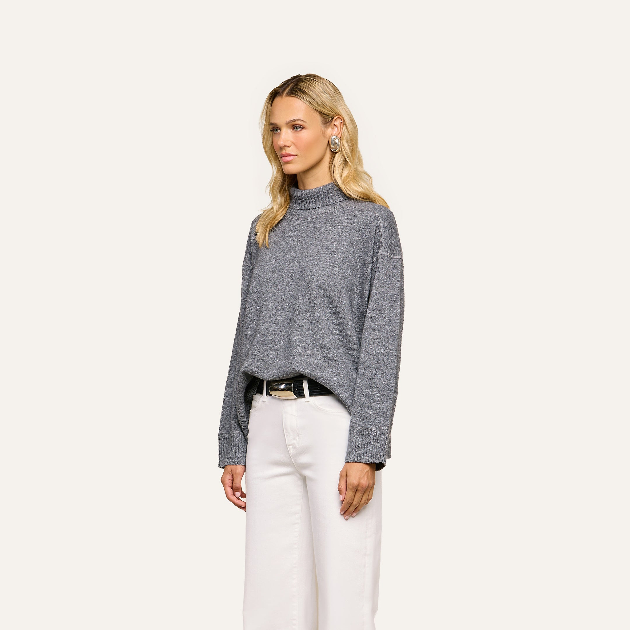 A woman with long blonde hair wears the Selwyn Oversized Turtleneck in heather grey with white pants, standing against a plain light background.