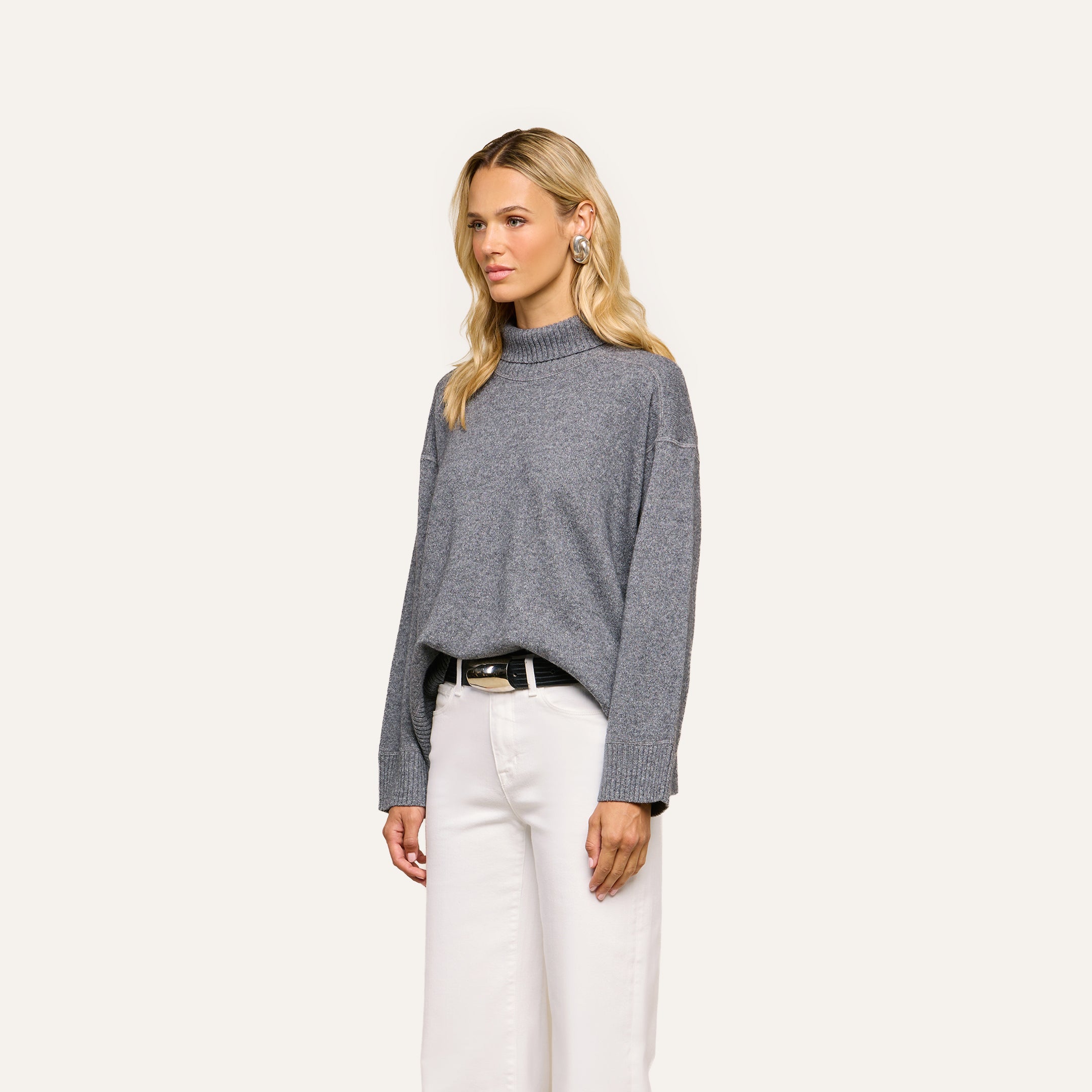 A woman with long blonde hair wears the Selwyn Oversized Turtleneck in heather grey with white pants, standing against a plain light background.