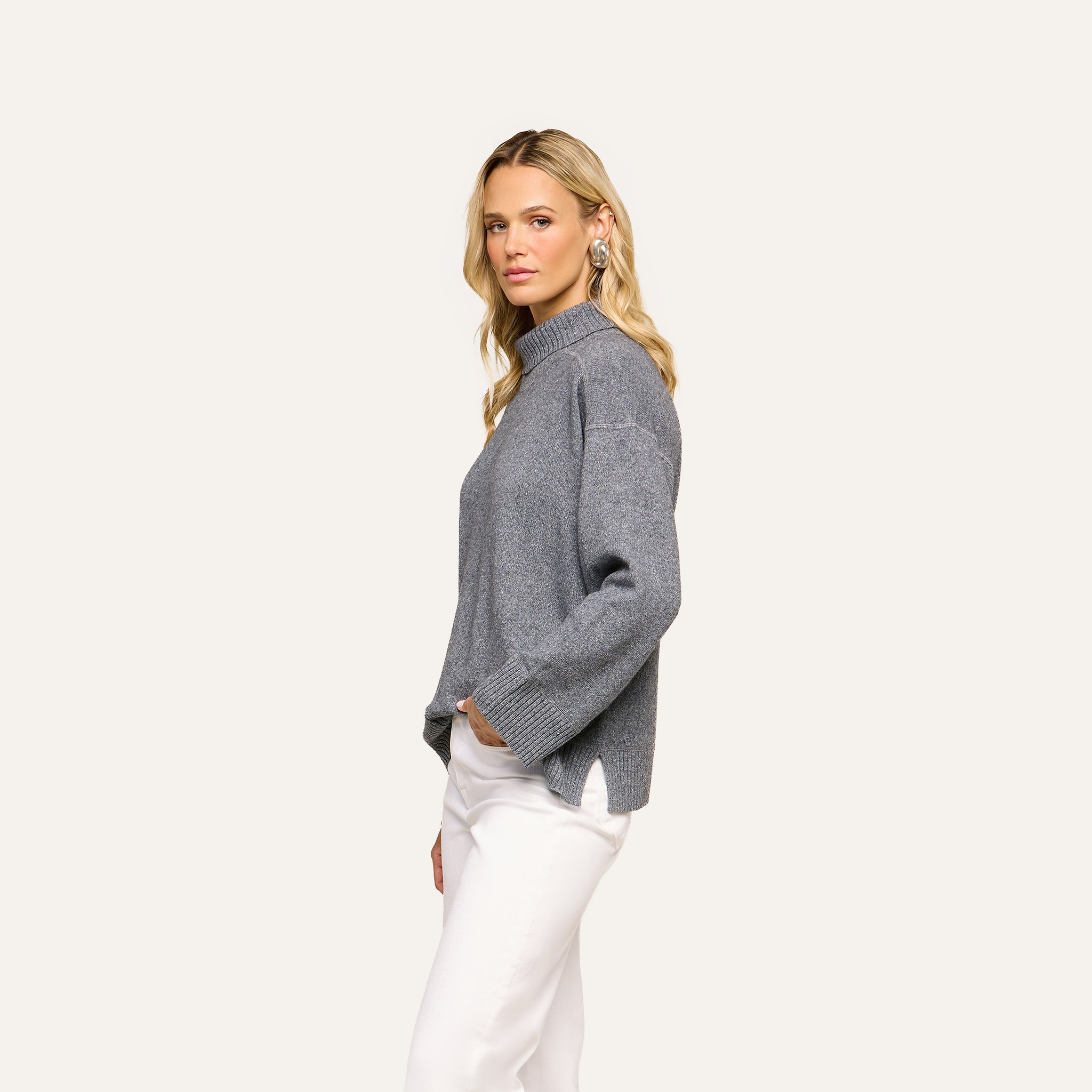 A woman with long blonde hair stands sideways against a plain light background, wearing the Selwyn Oversized Turtleneck in heather grey and white pants, with one hand in her pocket.