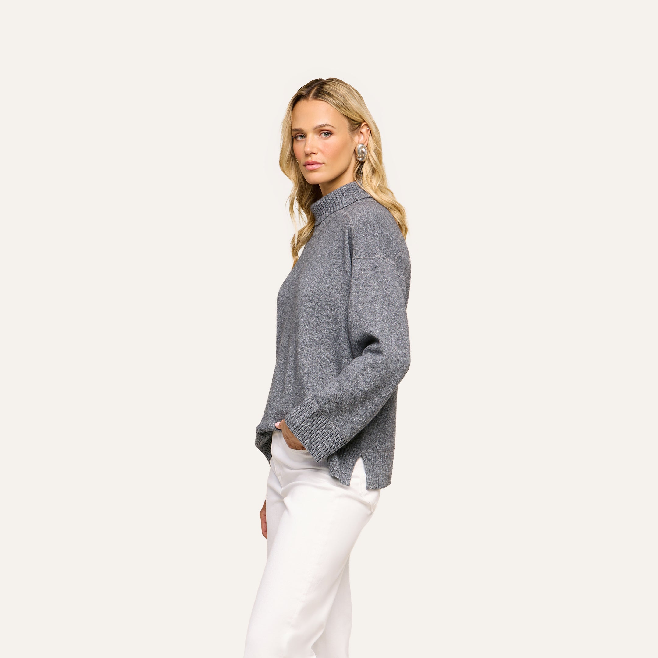 A woman with long blonde hair stands sideways against a plain light background, wearing the Selwyn Oversized Turtleneck in heather grey and white pants, with one hand in her pocket.