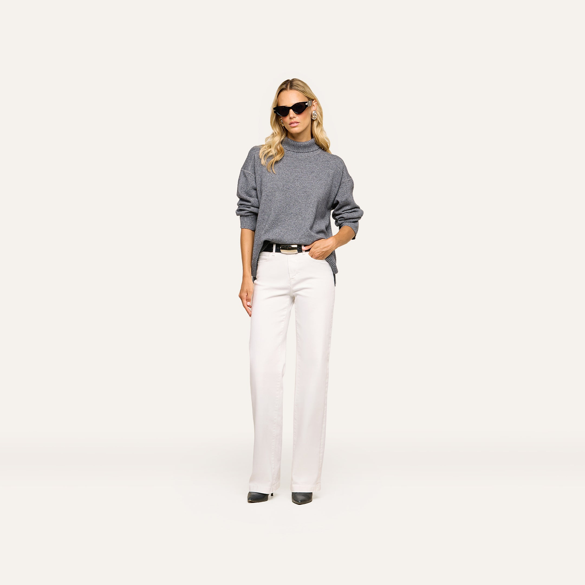 A woman wearing sunglasses, the Selwyn Oversized Turtleneck in heather grey wool cotton, white wide-leg pants, and black shoes stands against a plain, light background with one hand in her pocket.