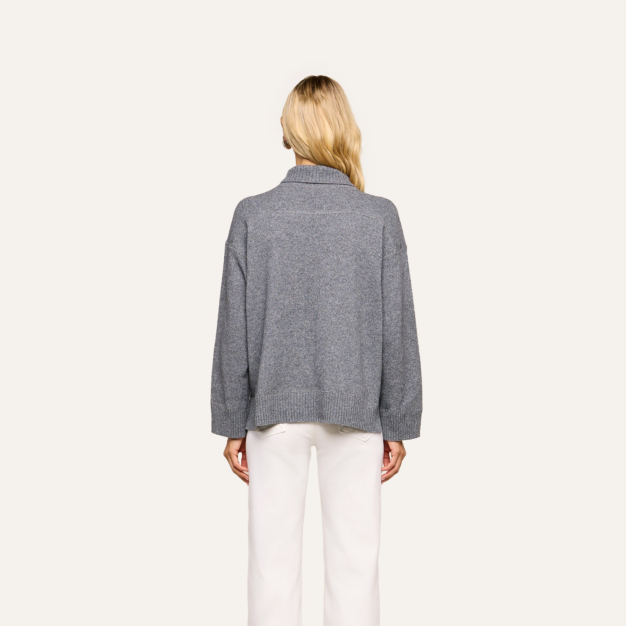 A person with long blonde hair, seen from behind, wears the Selwyn Oversized Turtleneck in heather grey paired with white pants, standing against a plain light background.