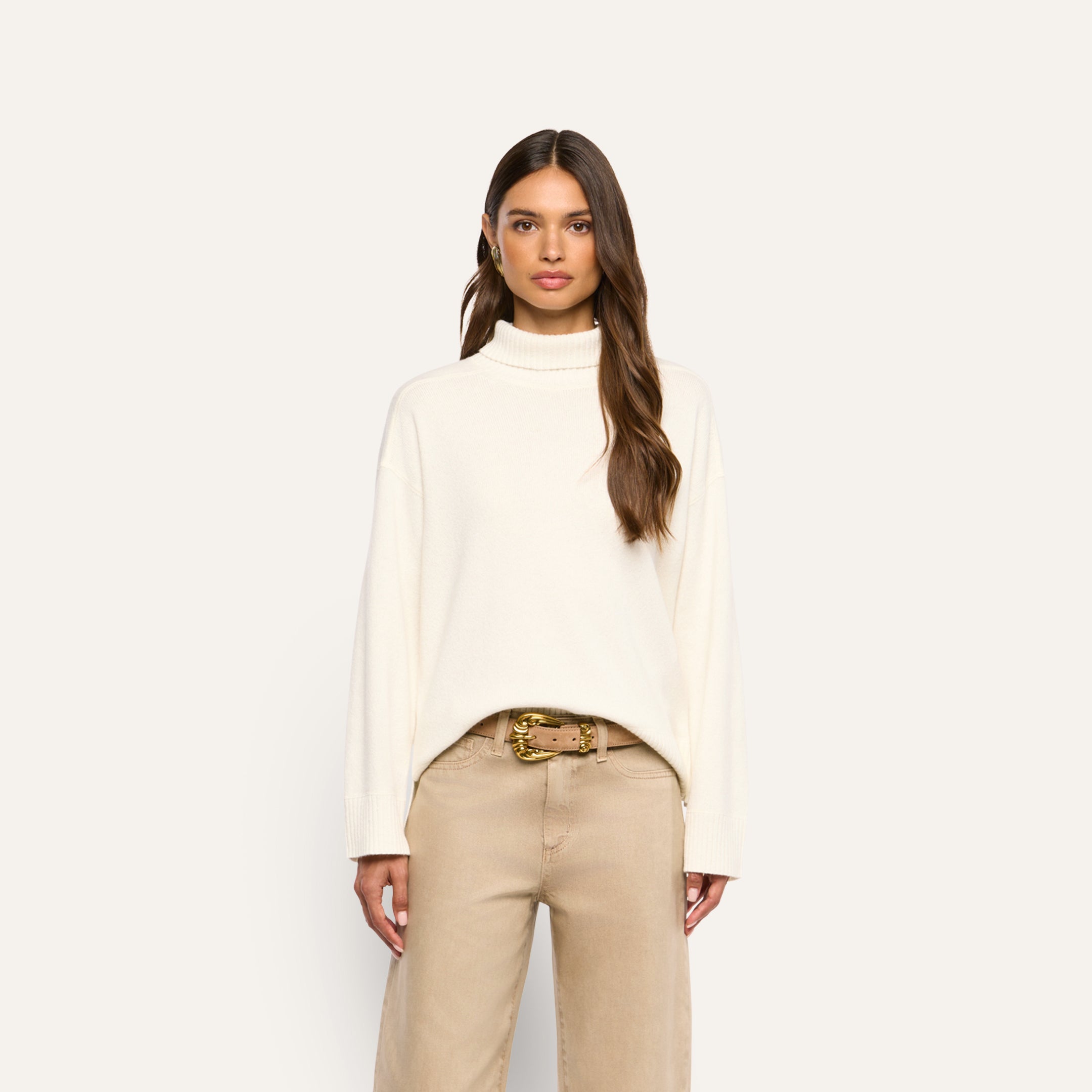 A woman wearing a Selwyn Oversized Turtleneck sweater tucked into tan trousers with a gold-buckled belt.