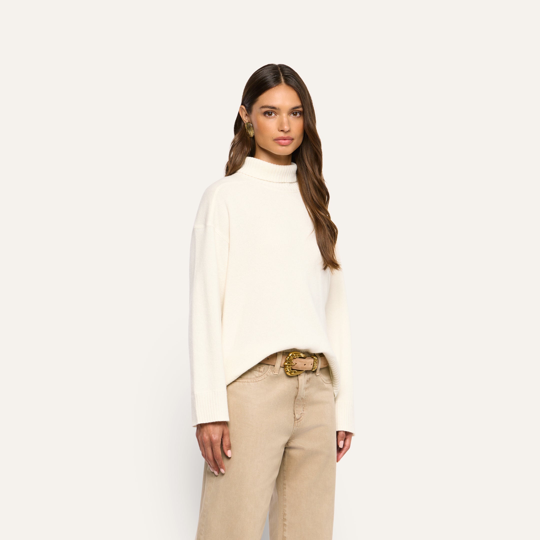 A woman wearing an oversized cream-colored merino wool Selwyn Oversized Turtleneck and tan jeans with a gold-buckled belt.
