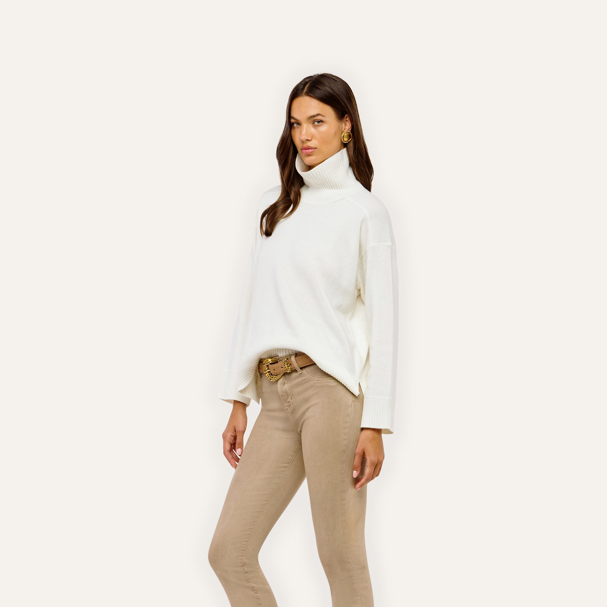 A person wears a Selwyn Oversized Turtleneck in premium cotton tucked into tan pants with a gold-buckle belt.