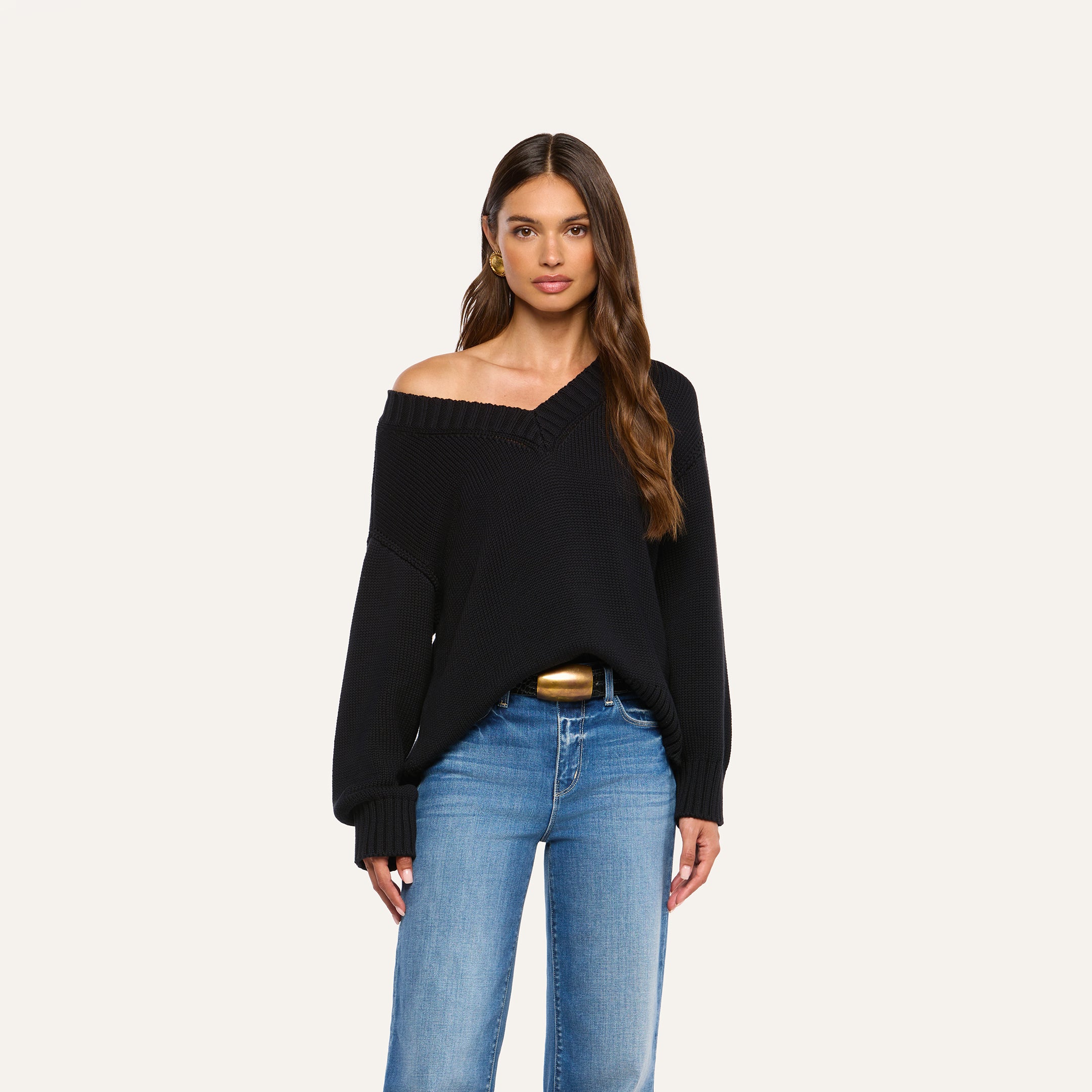 A woman with long brown hair wears the Paxton Oversized Pullover—a black, off-the-shoulder v-neck sweater—paired with blue jeans, standing against a plain, light background.