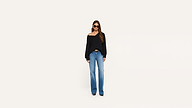 Woman wearing sunglasses, a black off-the-shoulder sweater, blue wide-leg jeans, and black shoes stands against a plain white background.