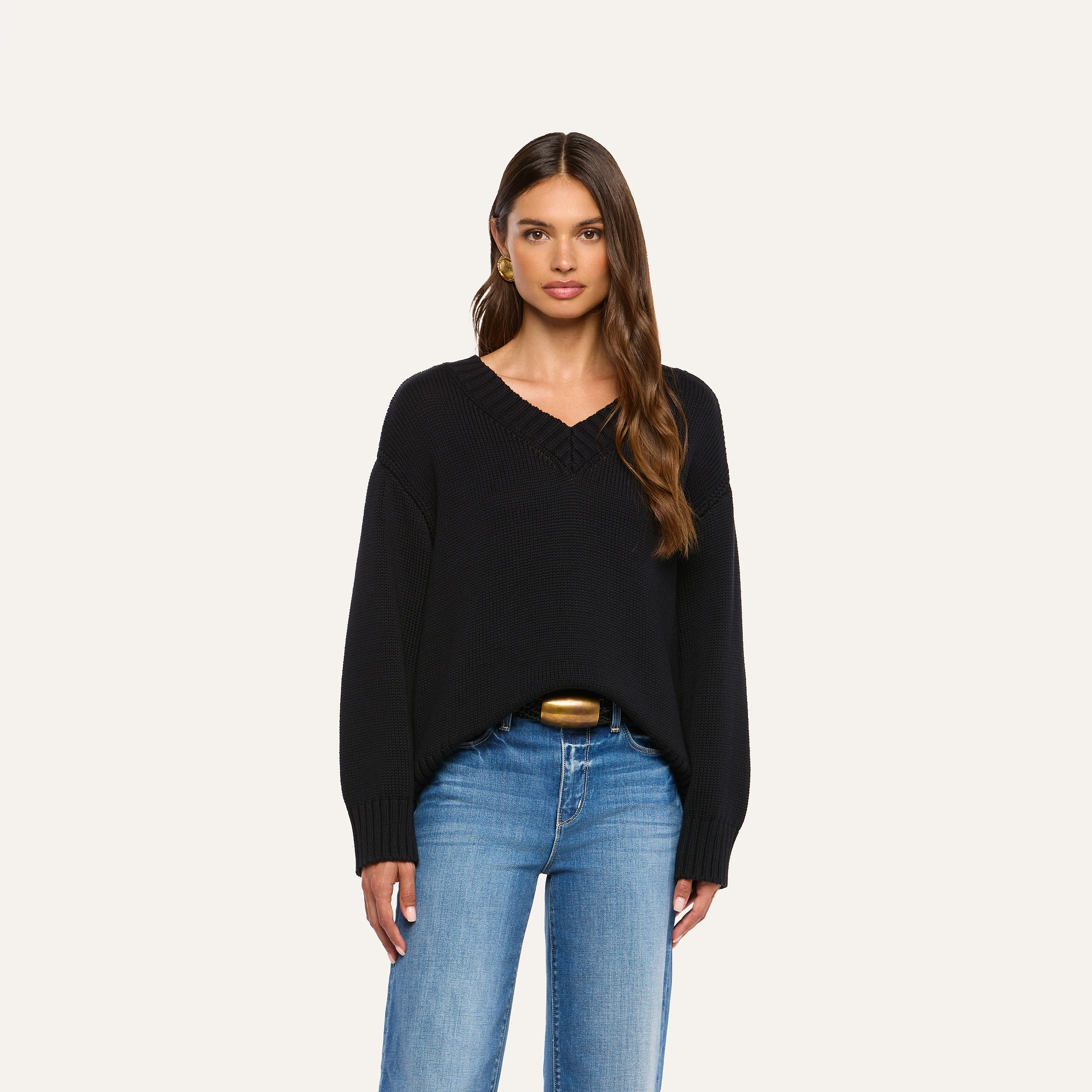 A woman with long brown hair wears the Paxton Oversized Pullover, a loose black v-neck sweater, paired with blue jeans. She stands facing forward against a plain light background.