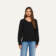 A woman with long brown hair wears the Paxton Oversized Pullover, a black cotton V-neck sweater, paired with blue jeans and stands against a plain light background.