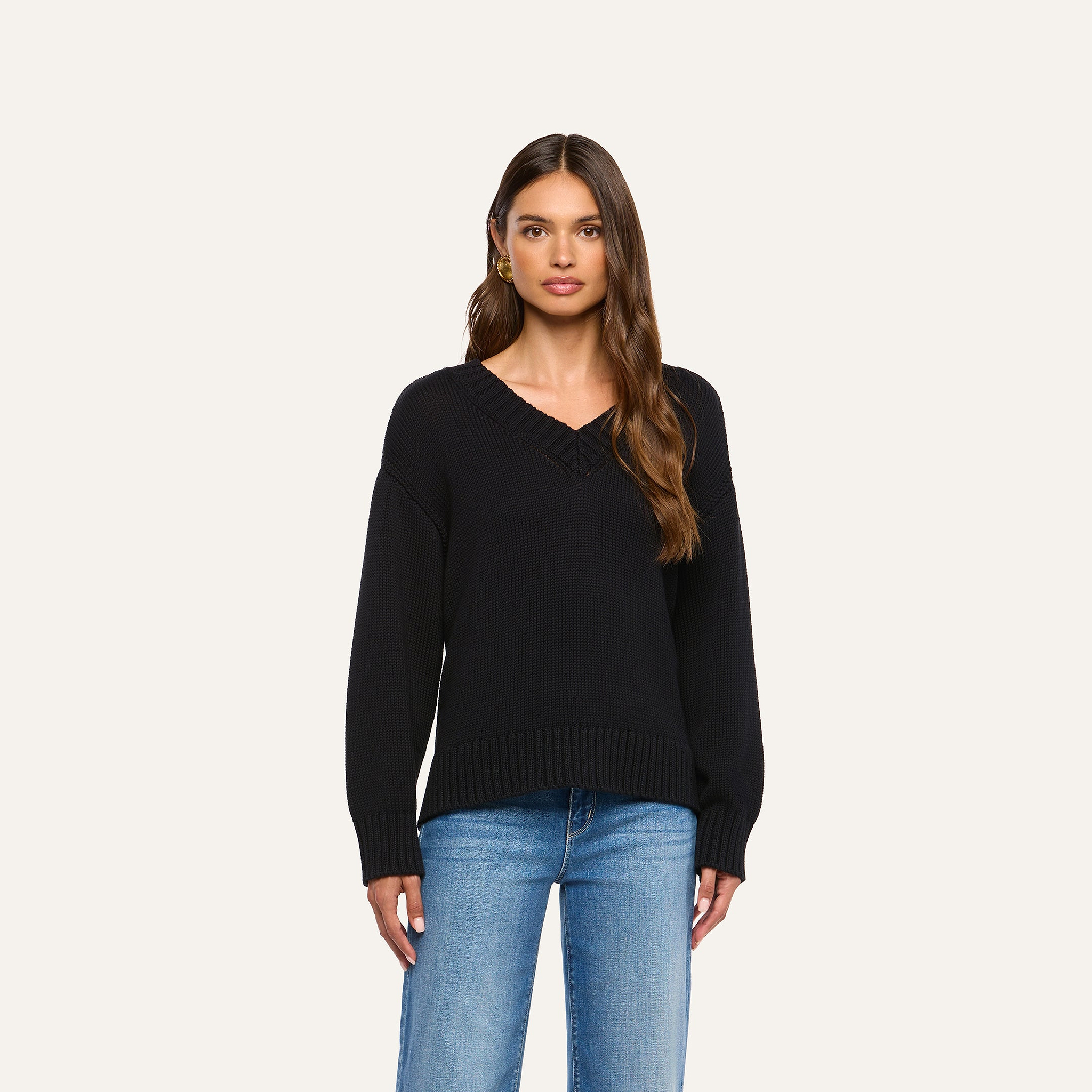 A woman with long brown hair wears the Paxton Oversized Pullover, a black cotton V-neck sweater, paired with blue jeans and stands against a plain light background.