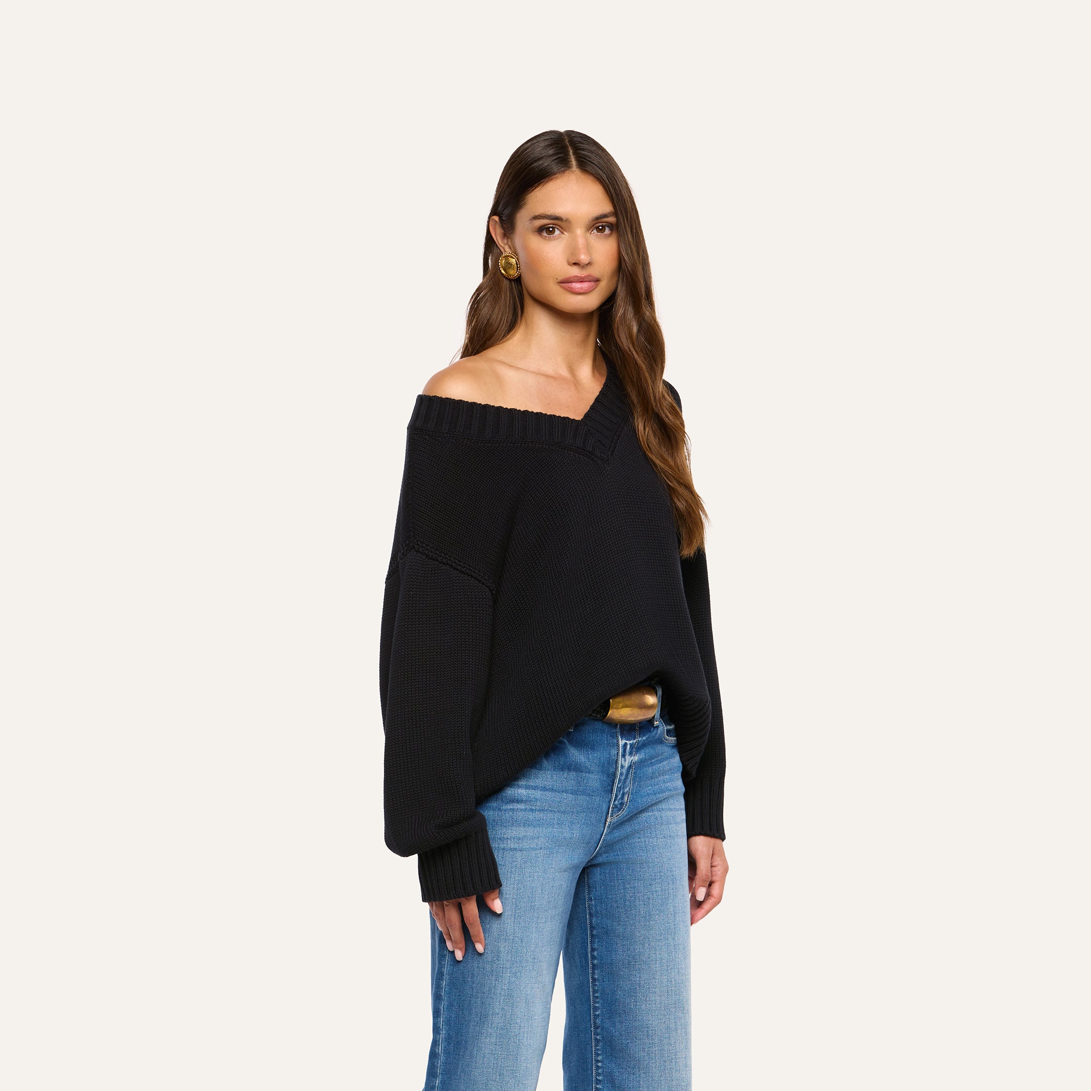 Woman with long brown hair wearing the Paxton Oversized Pullover, styled off-the-shoulder with blue jeans and gold earrings, stands against a plain light background, looking at the camera with a neutral expression.