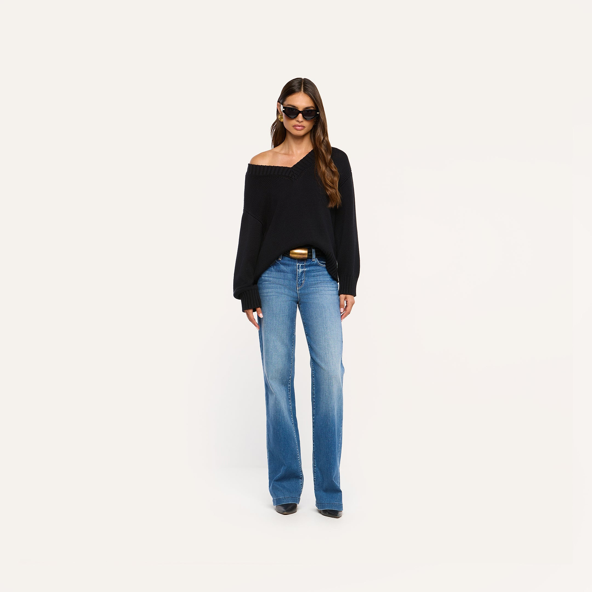 A woman wearing sunglasses, the Paxton Oversized Pullover in black off-the-shoulder style, blue high-waisted wide-leg jeans, and black shoes stands against a plain light background.
