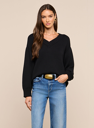 Wearing the Paxton Oversized Pullover in black, a woman with long brown hair and blue jeans is styled with gold hoop earrings and a gold-buckled belt, posed against a plain beige background.