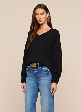 A woman with long brown hair wears the Paxton Oversized Pullover in black and blue jeans. She stands against a beige background, looking at the camera with a neutral expression.