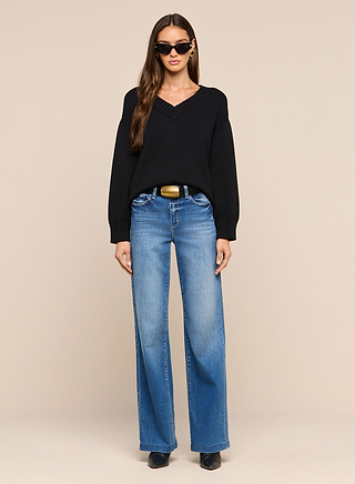 A woman stands facing forward against a plain beige background, wearing the Paxton Oversized Pullover in black, paired with blue wide-leg jeans, black boots, and sunglasses.