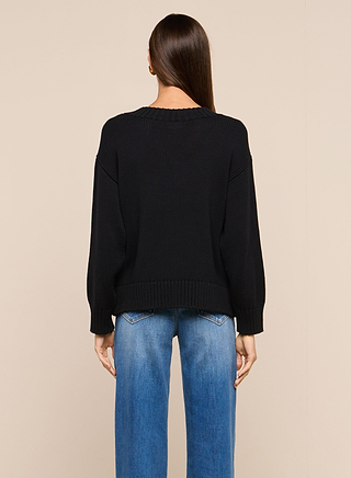 A person with long straight brown hair is seen from behind wearing the Paxton Oversized Pullover in black and blue jeans, standing against a plain beige background.