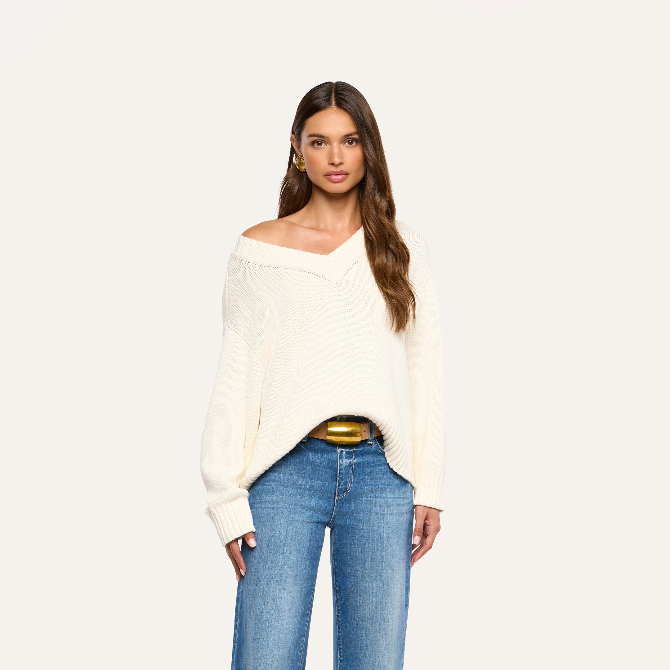 A woman with long brown hair wears the Paxton Oversized Pullover in ivory and blue jeans, standing against a plain light background.