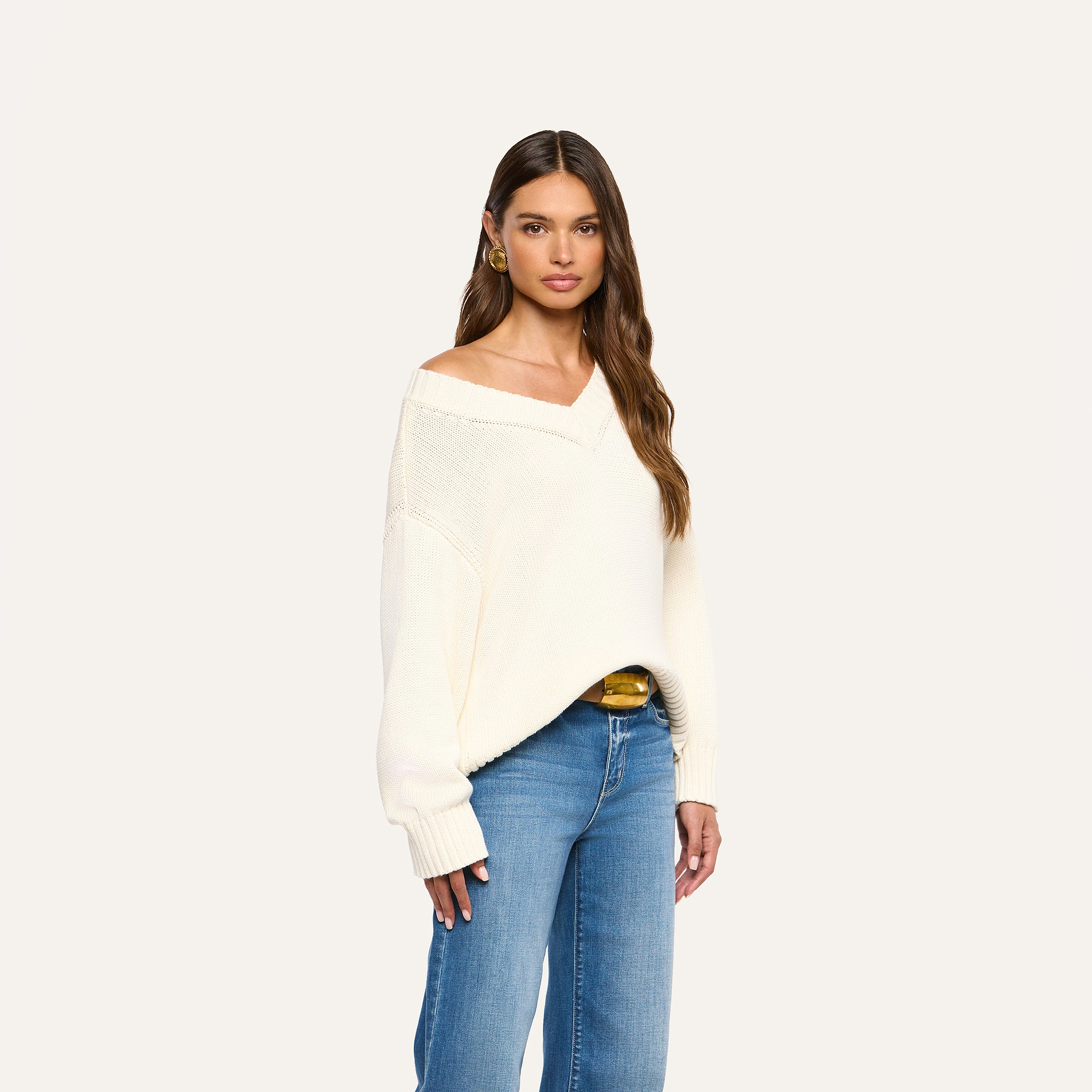 Woman with long brown hair wearing the Paxton Oversized Pullover in ivory cotton and blue jeans, posing against a plain light background.