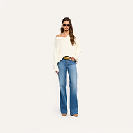 A woman stands against a plain background, wearing sunglasses, the Paxton Oversized Pullover in ivory, blue wide-leg jeans, brown pointed shoes, and hoop earrings. She has long wavy hair and holds a phone in her left hand.