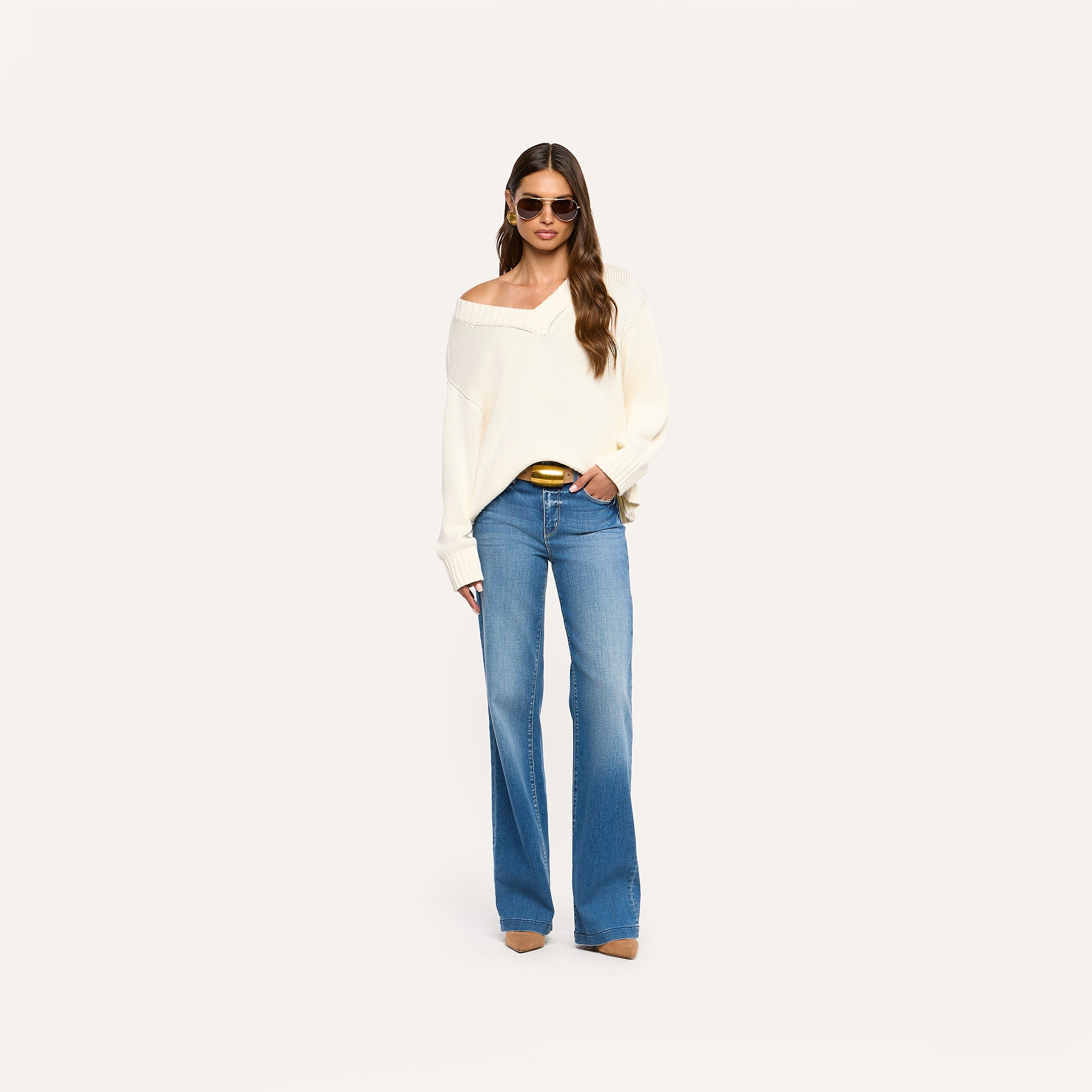 A woman stands against a plain background, wearing sunglasses, the Paxton Oversized Pullover in ivory, blue wide-leg jeans, brown pointed shoes, and hoop earrings. She has long wavy hair and holds a phone in her left hand.