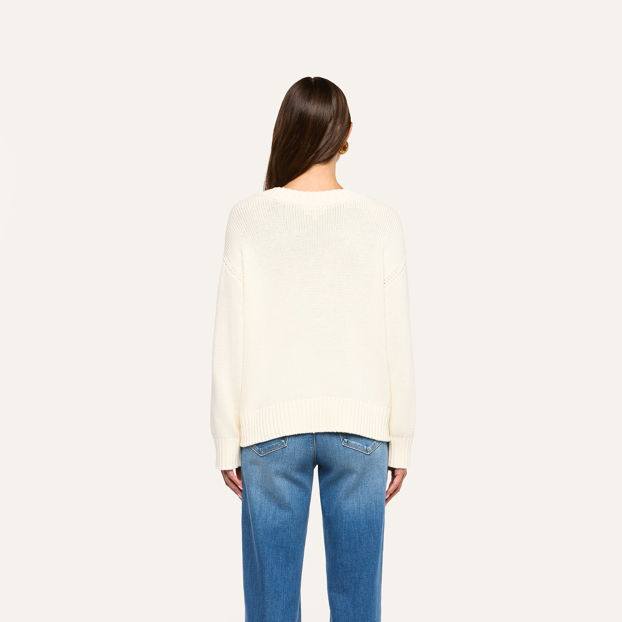 A person with long brown hair is facing away, wearing the Paxton Oversized Pullover in ivory and blue jeans, standing against a plain white background.