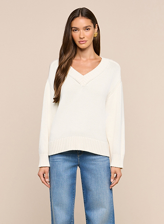 A woman wearing the Paxton Oversized Pullover, a cream-colored V-neck cotton sweater, with light blue jeans stands against a beige background, facing forward with a neutral expression.