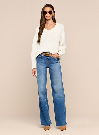 A woman wearing sunglasses, the Paxton Oversized Pullover in white, blue wide-leg jeans, and brown heels stands against a plain beige background with one hand in her pocket.