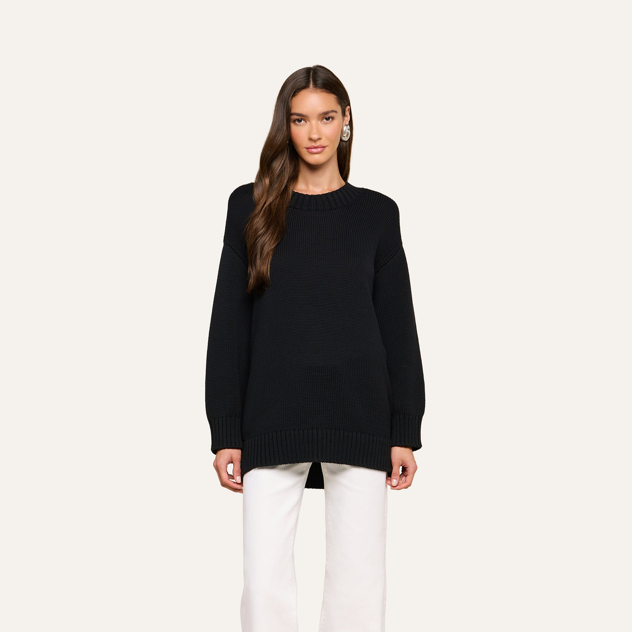 A woman with long brown hair wears the Loden Tunic Pullover in black paired with white pants, standing against a plain light background, facing forward with a neutral expression.