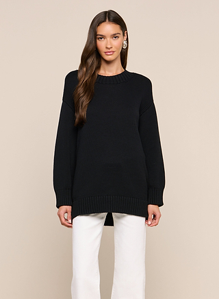 A woman with long brown hair wears the black Loden Pullover and white pants, standing against a plain beige background, looking at the camera with a neutral expression.